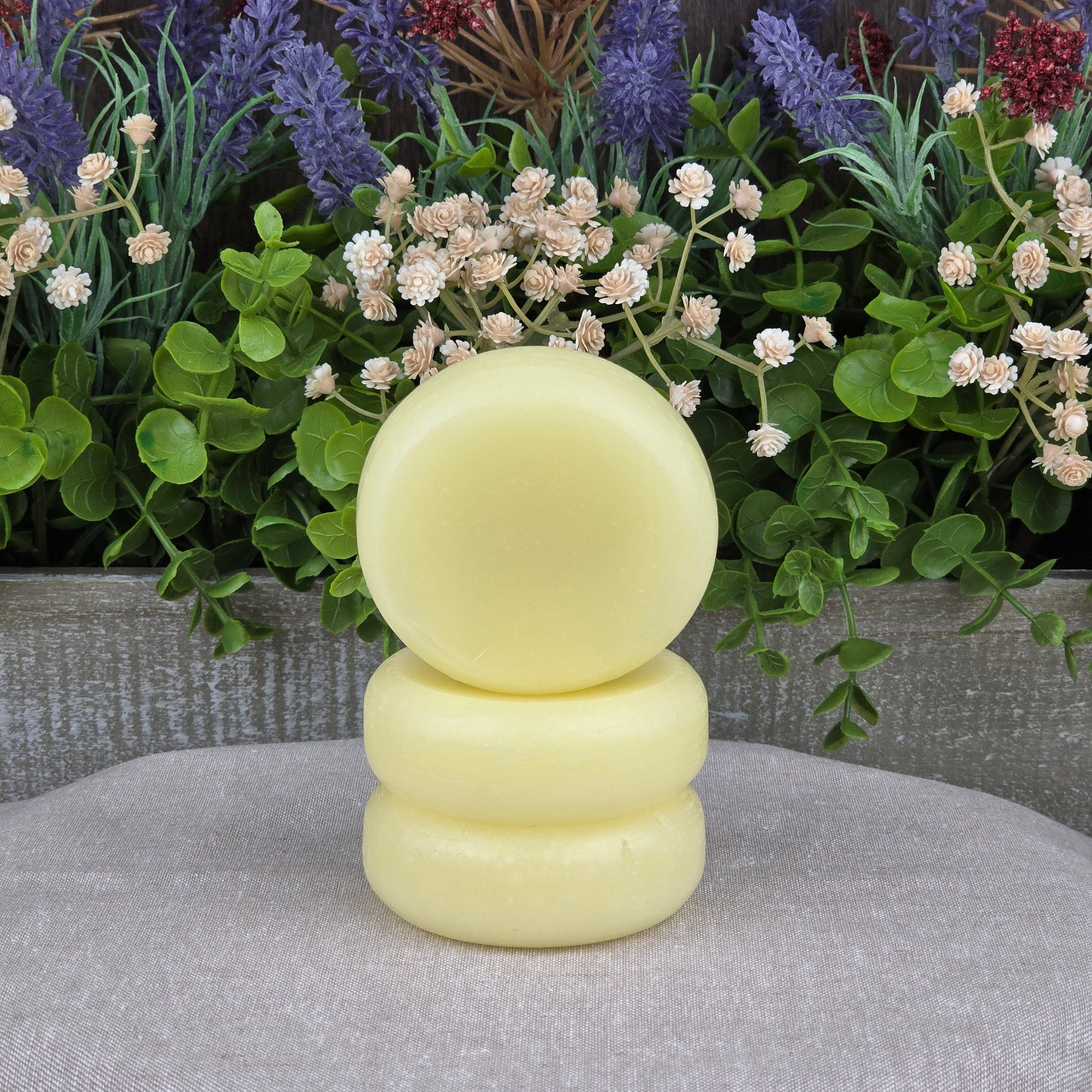 Three round, pale yellow Jarhead Aromatics Fragrance-Free Handmade Natural Conditioner Bars are stacked in front of lush artificial greenery and purple flowers, set against a wooden background with a light gray cloth underneath.