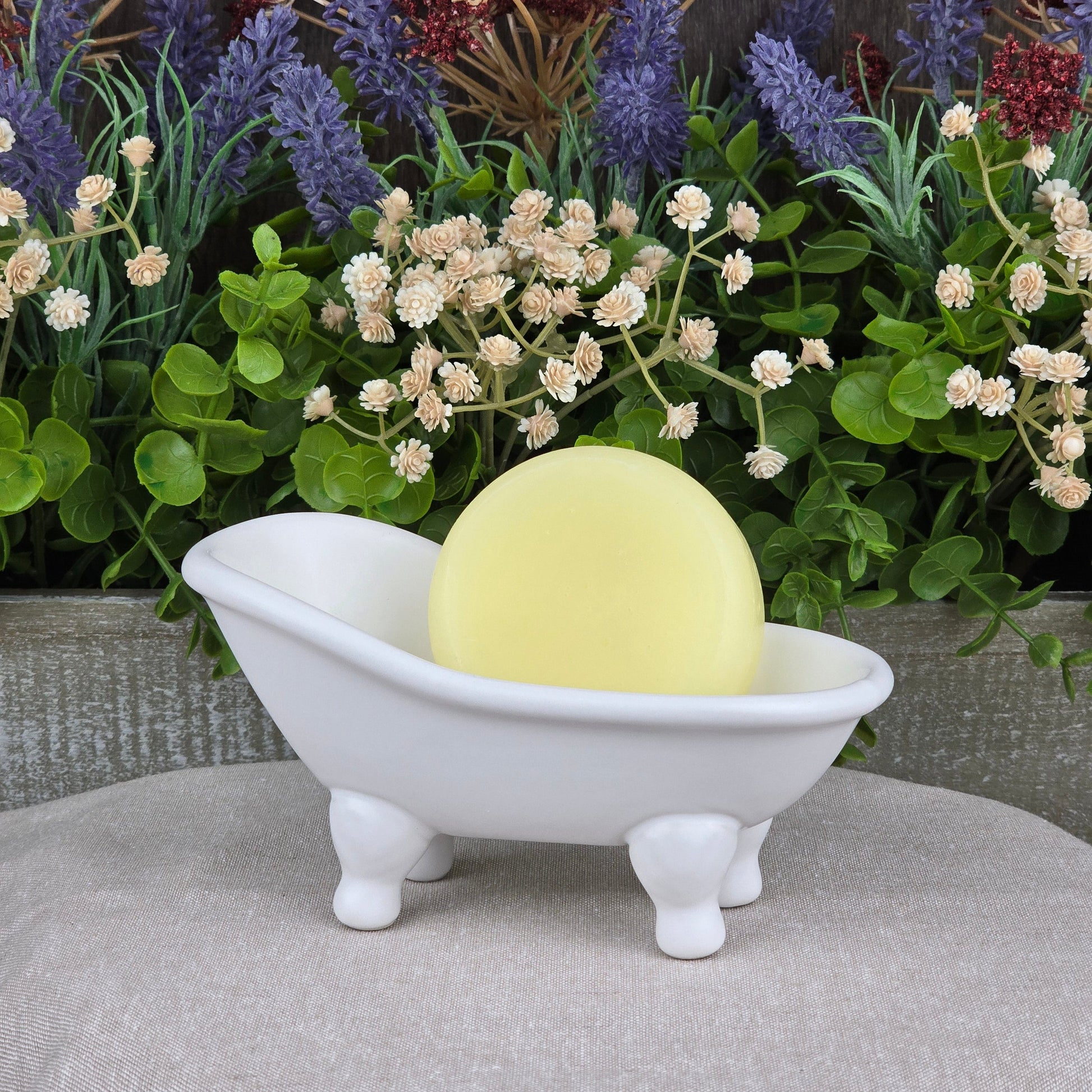 The Fragrance-Free Handmade Natural Conditioner Bar by Jarhead Aromatics—a round yellow bar—rests in a small, white, clawfoot tub-shaped dish with green plants and purple flowers in the background.