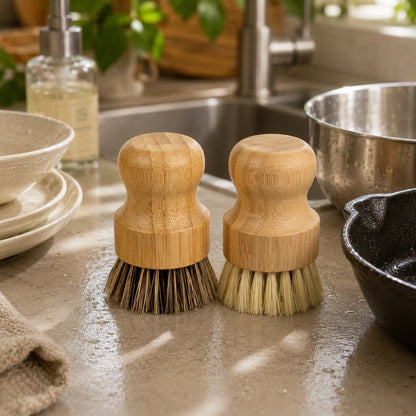 Two wooden-handled kitchen cleaning brushes from Jarhead Aromatics—a Bamboo Handle Palm & Sisal Fiber Scrubber Brush Set—rest on a wet countertop near dishes, a skillet, and the sink, with plants visible in the background.