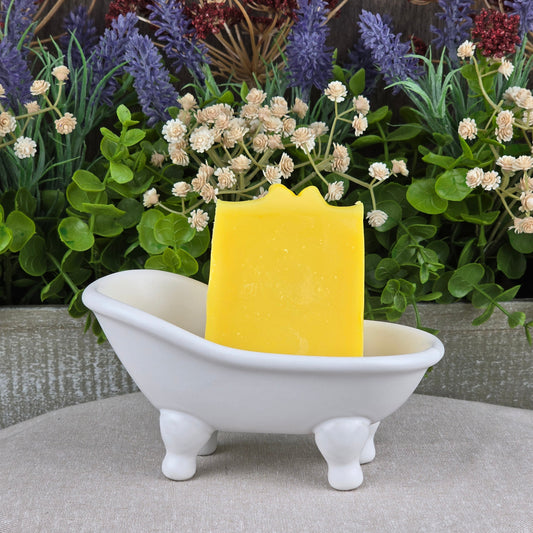 An Orange + Carrot Handmade Artisan Soap by Jarhead Aromatics stands upright in a small white ceramic bathtub, surrounded by green foliage and purple and white flowers.