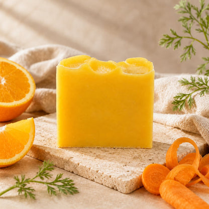 A bar of Jarhead Aromatics’ Orange + Carrot Handmade Natural Artisan Soap is displayed on a stone slab, surrounded by orange slices, carrot curls, and fresh green carrot tops, with a soft beige cloth in the background.