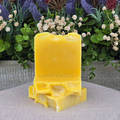 Two Orange + Carrot Handmade Natural Artisan Soap bars by Jarhead Aromatics are stacked in front of green leaves and colorful artificial flowers, creating a vibrant scene against a textured gray background.