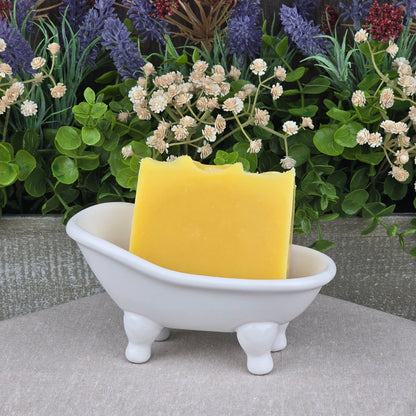 A yellow bar of Jarhead Aromatics’ Orange + Carrot Handmade Natural Artisan Soap rests in a white, bathtub-shaped dish, surrounded by artificial flowers and green foliage.