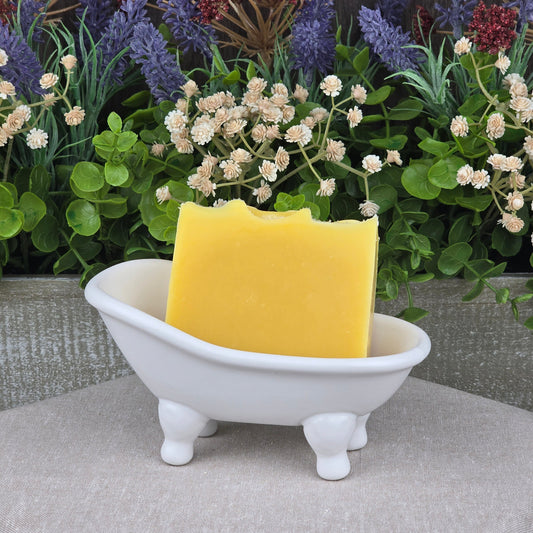 A yellow bar of Jarhead Aromatics’ Orange + Carrot Handmade Natural Artisan Soap rests in a white, bathtub-shaped dish, surrounded by artificial flowers and green foliage.