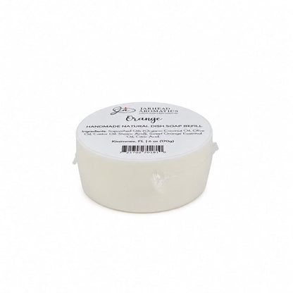 A round white handmade natural solid dish soap refill from Jarhead Aromatics, labeled with the scent “Orange,” ingredients, and a weight of 1.1 oz. This plant-based Orange Handmade Natural Solid Dish Soap | Citrus Kitchen Cleaning Bar with Essential Oils is wrapped in plastic and features a white top label displaying branding details.