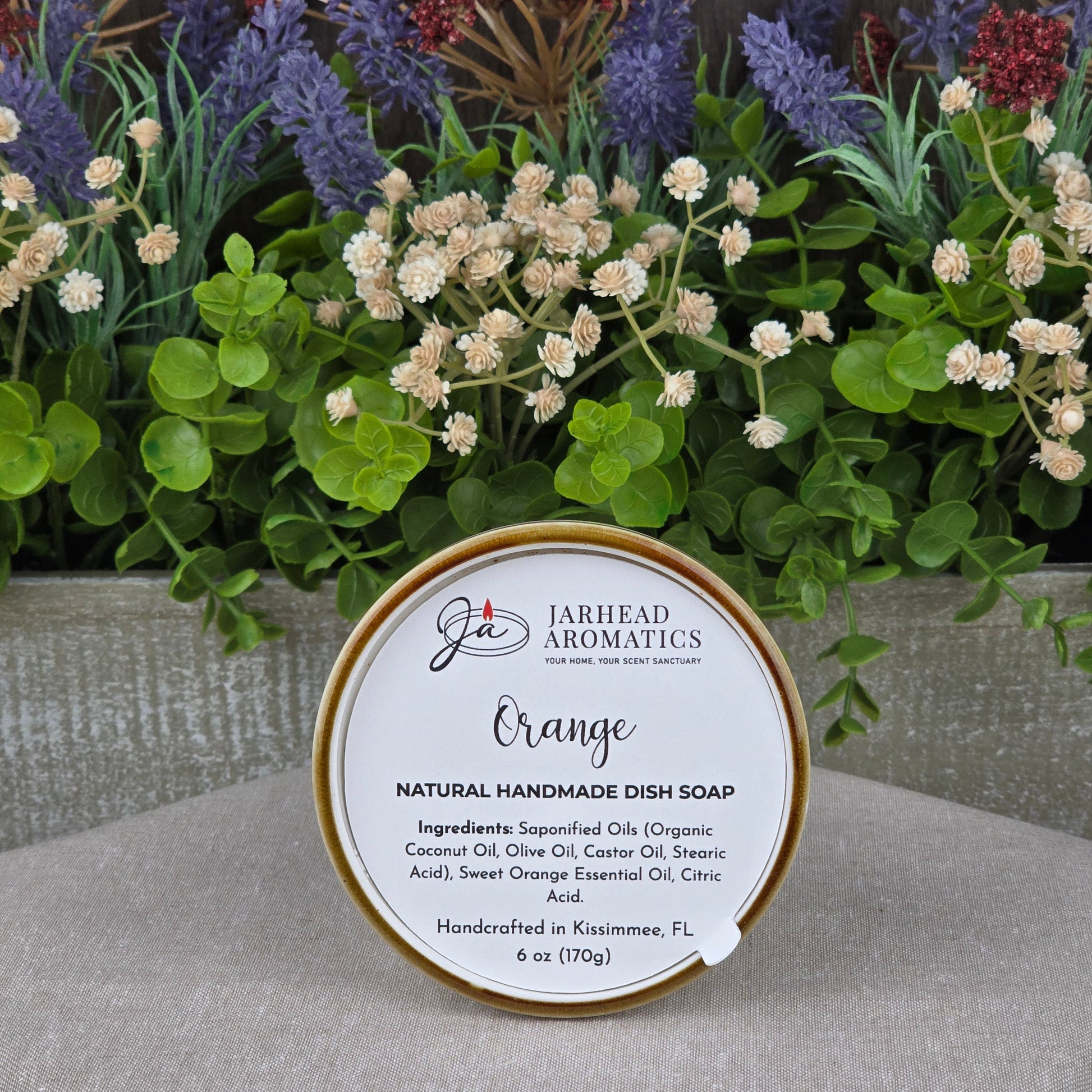 A round tin of Jarhead Aromatics Orange Handmade Natural Solid Dish Soap rests on a stone surface, its label visible. Colorful artificial flowers and greenery create a charming background.