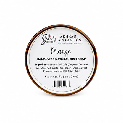 A round tin labeled "Jarhead Aromatics Orange Handmade Natural Solid Dish Soap | Citrus Kitchen Cleaning Bar with Essential Oils" displays its ingredients and a weight of 6 oz (170g) against a white background with black text, showcasing this plant-based cleaning bar as a natural solid dish soap.