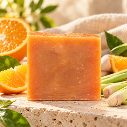 A rectangular Orange + Lemongrass Natural Handmade Goat Milk Soap bar placed on a stone surface surrounded by orange slices and stalks.