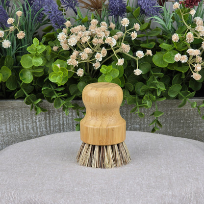 The Jarhead Aromatics Bamboo Handle Palm Fiber Scrubber Brush rests on a light gray surface, accented by green leaves and flowers—an eco-friendly, heavy-duty cleaning brush ideal for sustainable homes.