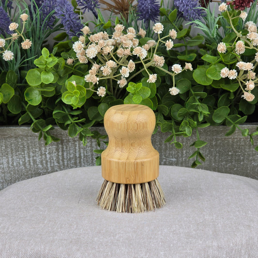 The Jarhead Aromatics Bamboo Handle Palm Fiber Scrubber Brush rests on a light gray surface, accented by green leaves and flowers—an eco-friendly, heavy-duty cleaning brush ideal for sustainable homes.