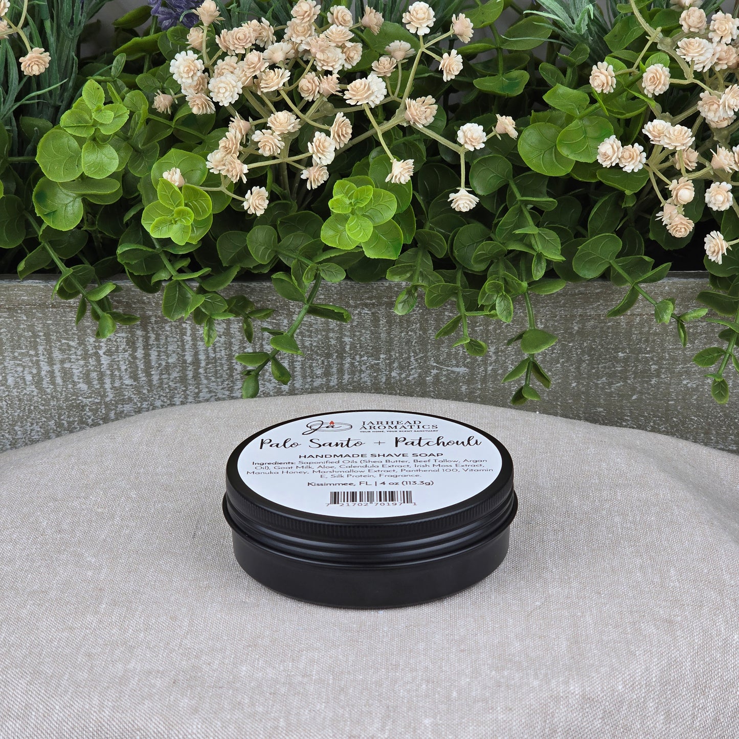 A black tin of Jarhead Aromatics Palo Santo + Patchouli Handmade Shave Soap rests on a light gray cushion, with a wooden planter of green leaves and cream flowers in the background. The natural tallow & goat milk soap blends artisan care with quality ingredients.