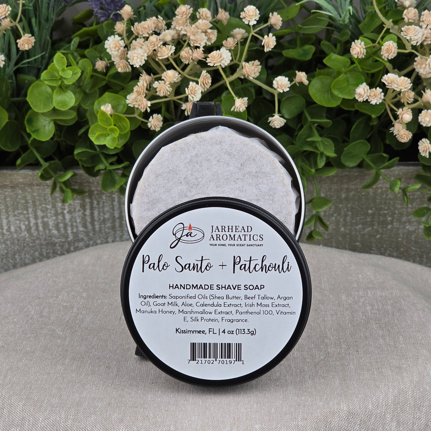 A round tin of Jarhead Aromatics Palo Santo + Patchouli Handmade Shave Soap rests on a gray surface, bordered by green leaves and white flowers. The label highlights its natural tallow and goat milk ingredients.