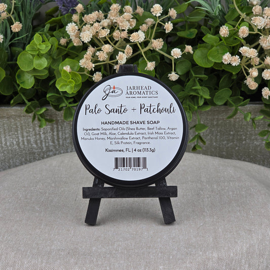 A round Jarhead Aromatics Palo Santo + Patchouli Handmade Shave Soap sits on a small black easel, set against green plants and a gray textured background. The container displays ingredients and product details.