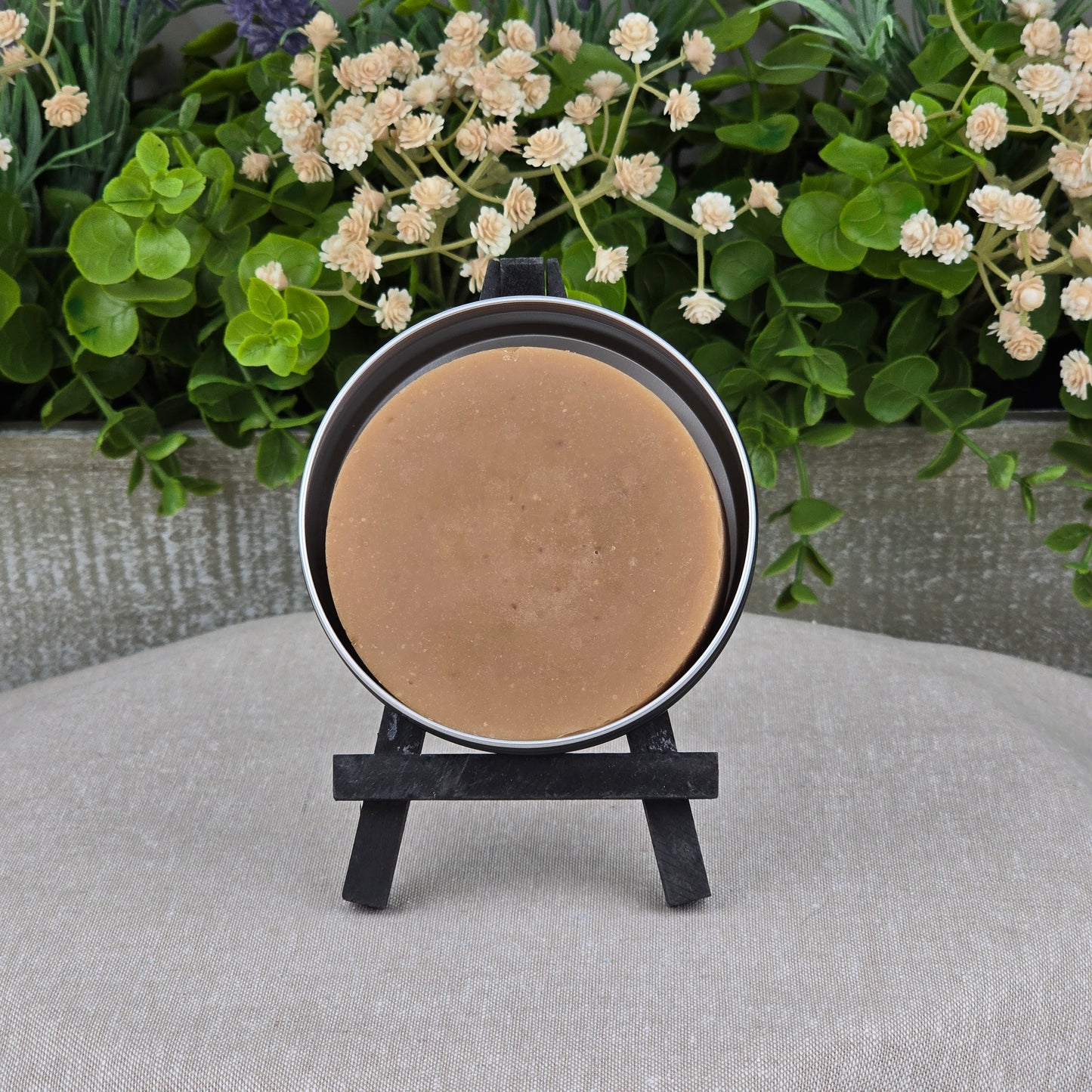 A round brown bar of Palo Santo + Patchouli Handmade Shave Soap by Jarhead Aromatics, made with natural tallow & goat milk, is displayed upright in a metal tin on a black easel, with green leaves and white flowers in the background.