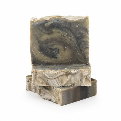 Three rectangular bars of marbled, handcrafted soap in shades of gray, olive, and cream are stacked on a white background. The rustic, swirled patterns and wavy edges showcase the artisanal quality of each bar from Jarhead Aromatics. Among these distinctive creations is our Palo Santo + Patchouli Handmade Artisan Soap | Smoky Woodsy Charcoal Soap—a unique blend of earthy aromas and striking design, ideal for thoughtful gifting or everyday indulgence.