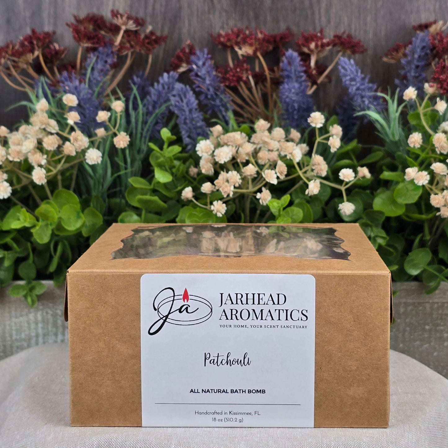 A box labeled Jarhead Aromatics contains the Patchouli Bath Bomb - All Natural, infused with soothing patchouli. Its beautifully displayed against lavender, white flowers, and greenery.