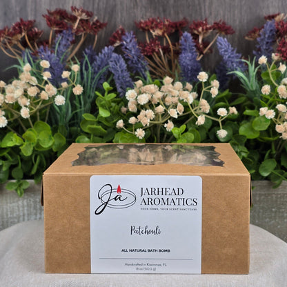 A box labeled Jarhead Aromatics contains the Patchouli Bath Bomb - All Natural, infused with soothing patchouli. Its beautifully displayed against lavender, white flowers, and greenery.