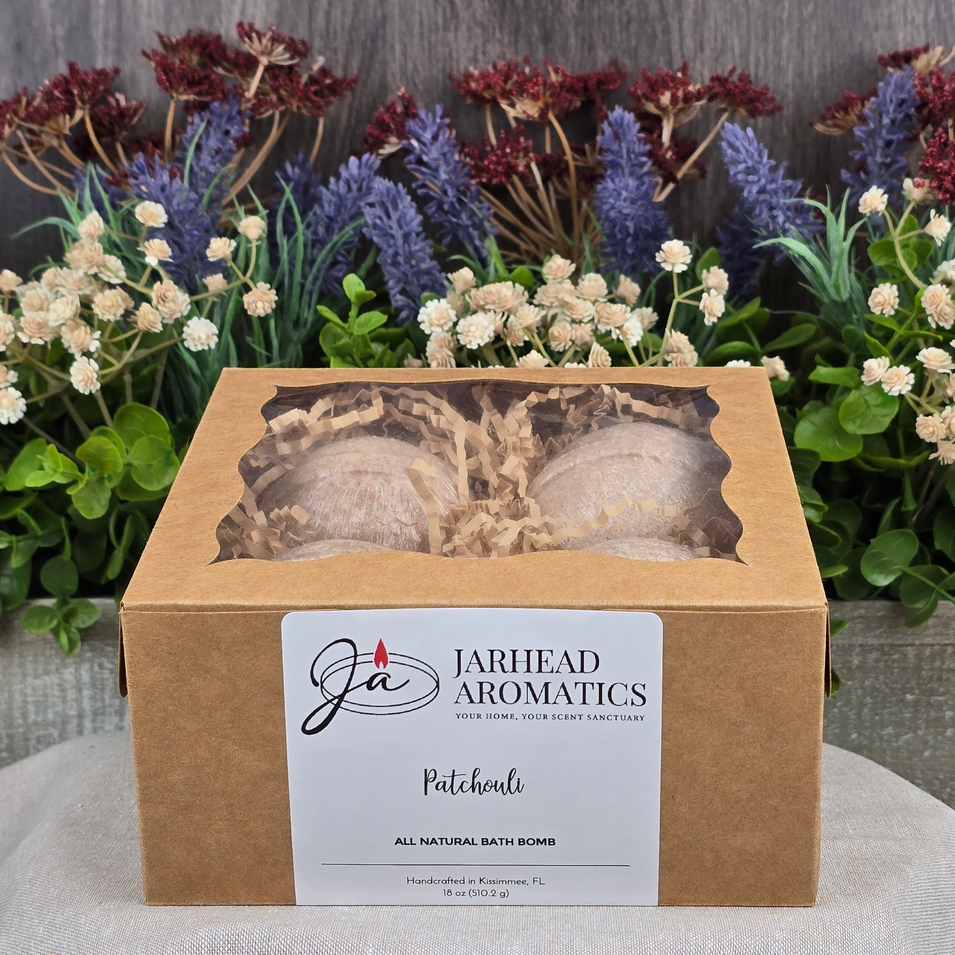 A cardboard box of Jarhead Aromatics Patchouli Bath Bomb - All Natural, featuring a refreshing scent, displayed with artificial flowers in white, red, and purple hues. The label highlights its made from natural ingredients and handcrafted in Kissimmee, FL.