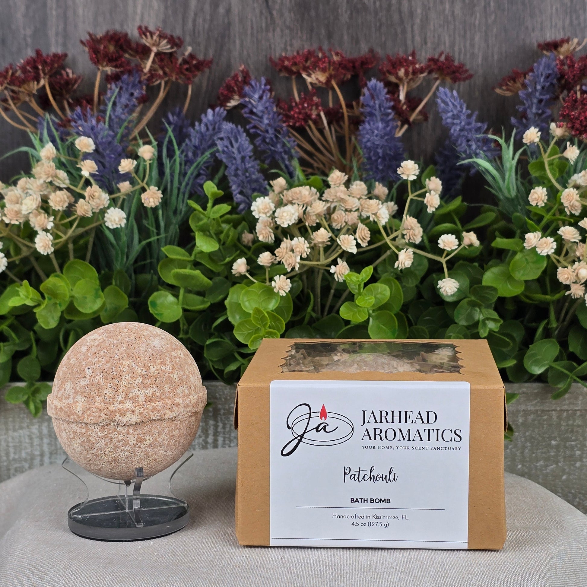 Displayed on a stand beside its box, the round, handmade Patchouli Bath Bomb - All Natural by Jarhead Aromatics is set against a gray backdrop. Artificial plants with green leaves and colorful red, purple, and white flowers enhance the natural vibe.