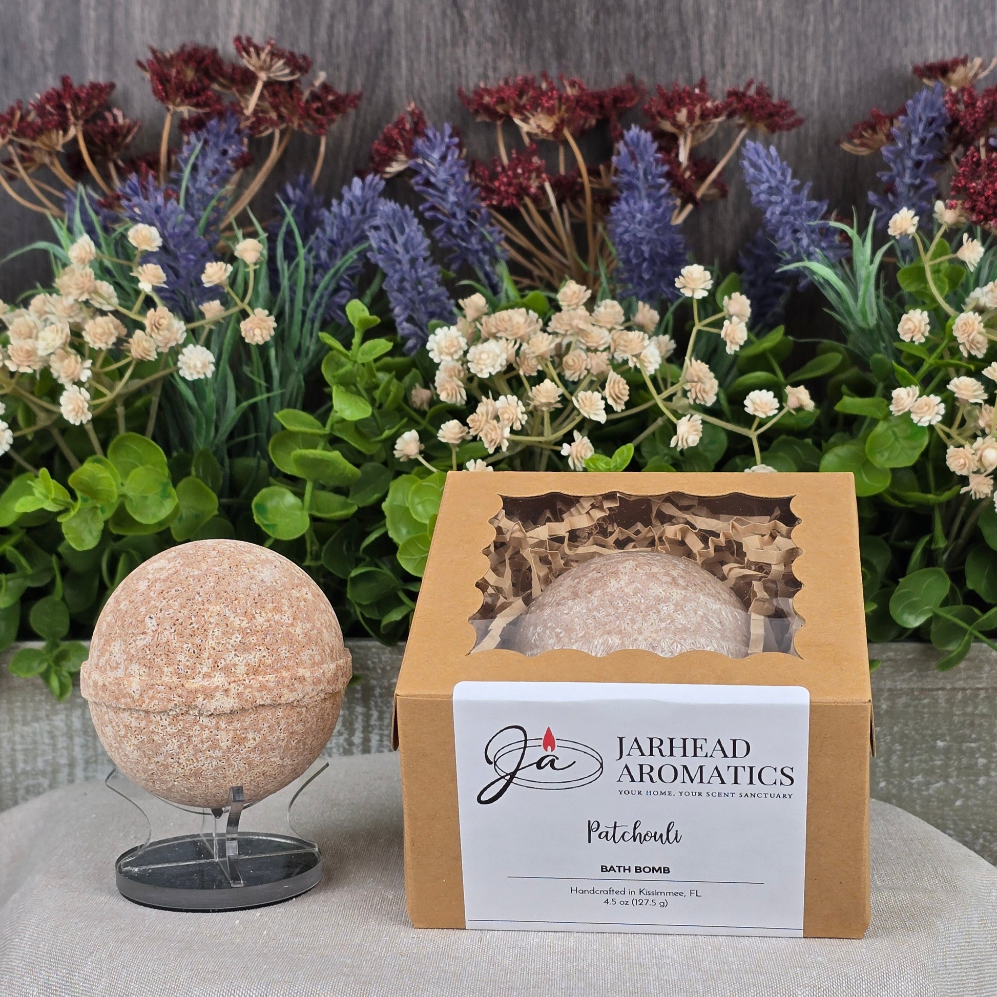 A speckled beige Patchouli Bath Bomb from Jarhead Aromatics, crafted with natural ingredients, sits alongside a branded box labeled Patchouli. In the background, artificial foliage adds lavender, white, and burgundy floral accents.