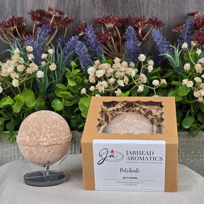 A speckled beige Patchouli Bath Bomb from Jarhead Aromatics, crafted with natural ingredients, sits alongside a branded box labeled Patchouli. In the background, artificial foliage adds lavender, white, and burgundy floral accents.