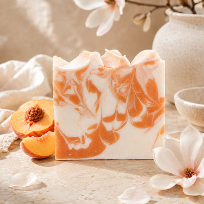 A decorative bar of Jarhead Aromatics Peach + Magnolia Handmade Coconut Milk Artisan Soap, marbled in orange and white, rests on a light surface surrounded by peach slices, white flowers, and a ceramic vase. This juicy peach and floral magnolia soap is handcrafted with nourishing coconut milk for a sumptuous experience.