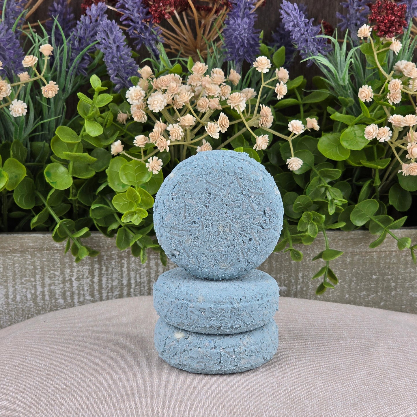Three round, blue Jarhead Aromatics Peppermint Handmade Shampoo & Conditioner Bars sit on a beige surface amid green foliage and blooms—an elegant, all-natural option for zero-waste hair care.