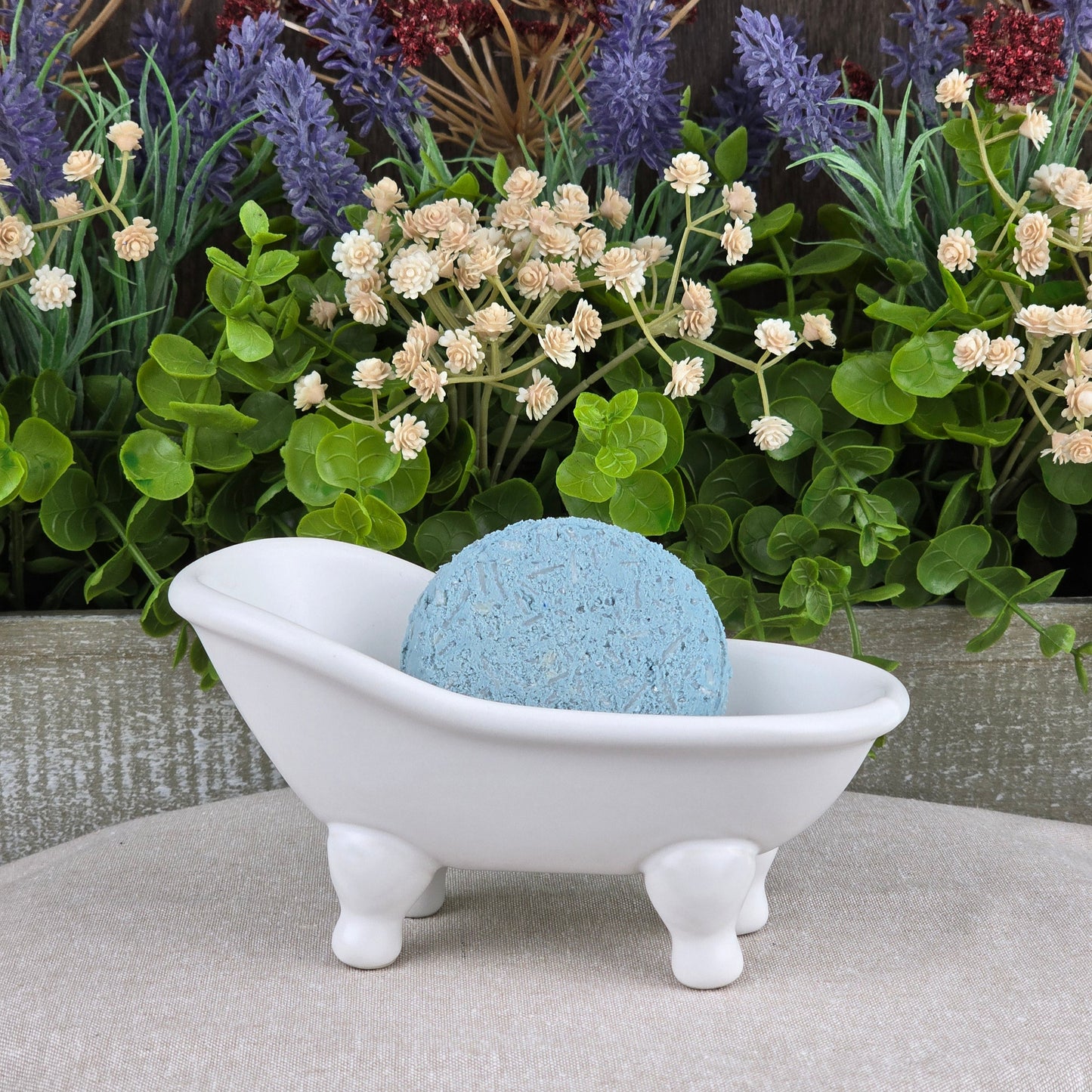 A blue round bath bomb rests in a white ceramic tub, surrounded by green leaves and faux flowers—the ideal match for Jarhead Aromatics’ Peppermint Handmade Shampoo & Conditioner Bar, supporting your all-natural, zero-waste hair care routine.