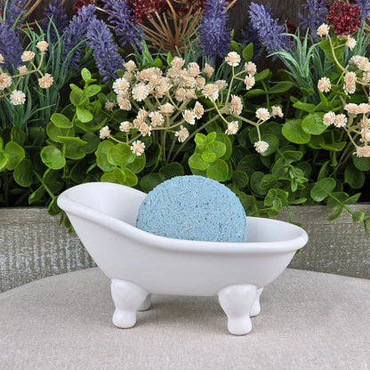 A blue round bath bomb rests in a white ceramic tub, surrounded by green leaves and faux flowers—the ideal match for Jarhead Aromatics’ Peppermint Handmade Shampoo & Conditioner Bar, supporting your all-natural, zero-waste hair care routine.