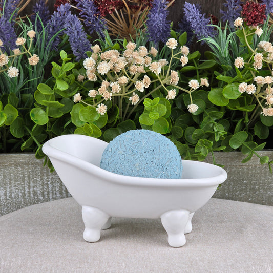 A blue round bath bomb rests in a white ceramic tub, surrounded by green leaves and faux flowers—the ideal match for Jarhead Aromatics’ Peppermint Handmade Shampoo & Conditioner Bar, supporting your all-natural, zero-waste hair care routine.