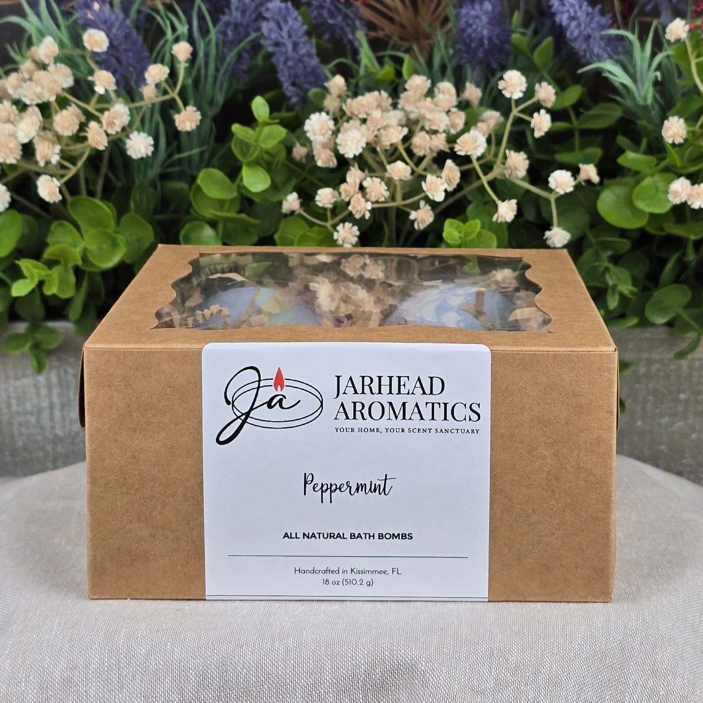 A brown box labeled Jarhead Aromatics Peppermint Bath Bomb - All Natural sits on a white surface, with artificial flowers and greenery in the background, highlighting the bath bomb’s natural appeal.