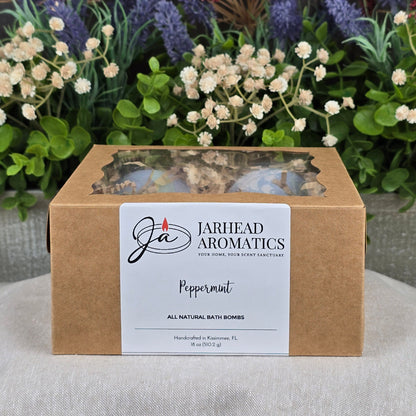 A brown box labeled Jarhead Aromatics Peppermint Bath Bomb - All Natural sits on a white surface, with artificial flowers and greenery in the background, highlighting the bath bomb’s natural appeal.