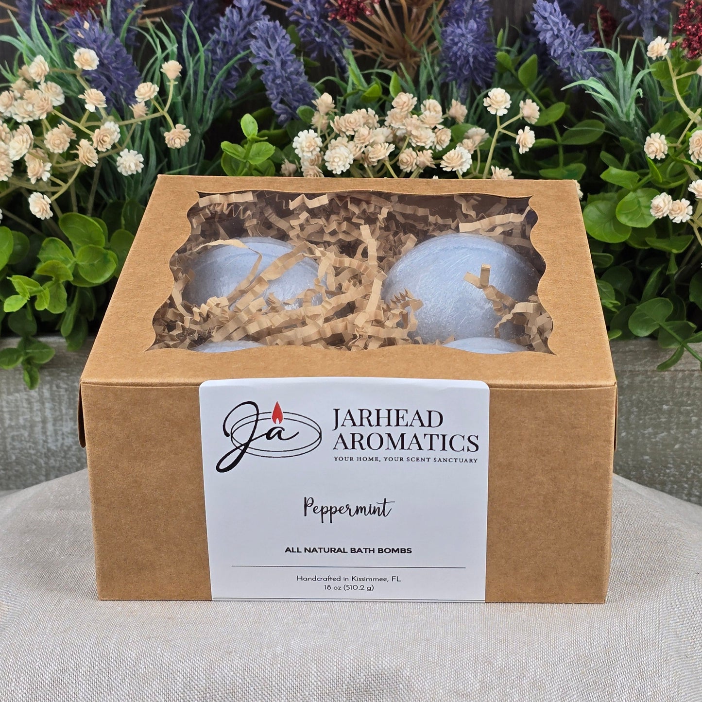 A cardboard box of Jarhead Aromatics Peppermint Bath Bomb - All Natural rests on a gray surface, surrounded by artificial greenery and purple flowers. A clear window on the box shows two blue handmade bath bombs inside.