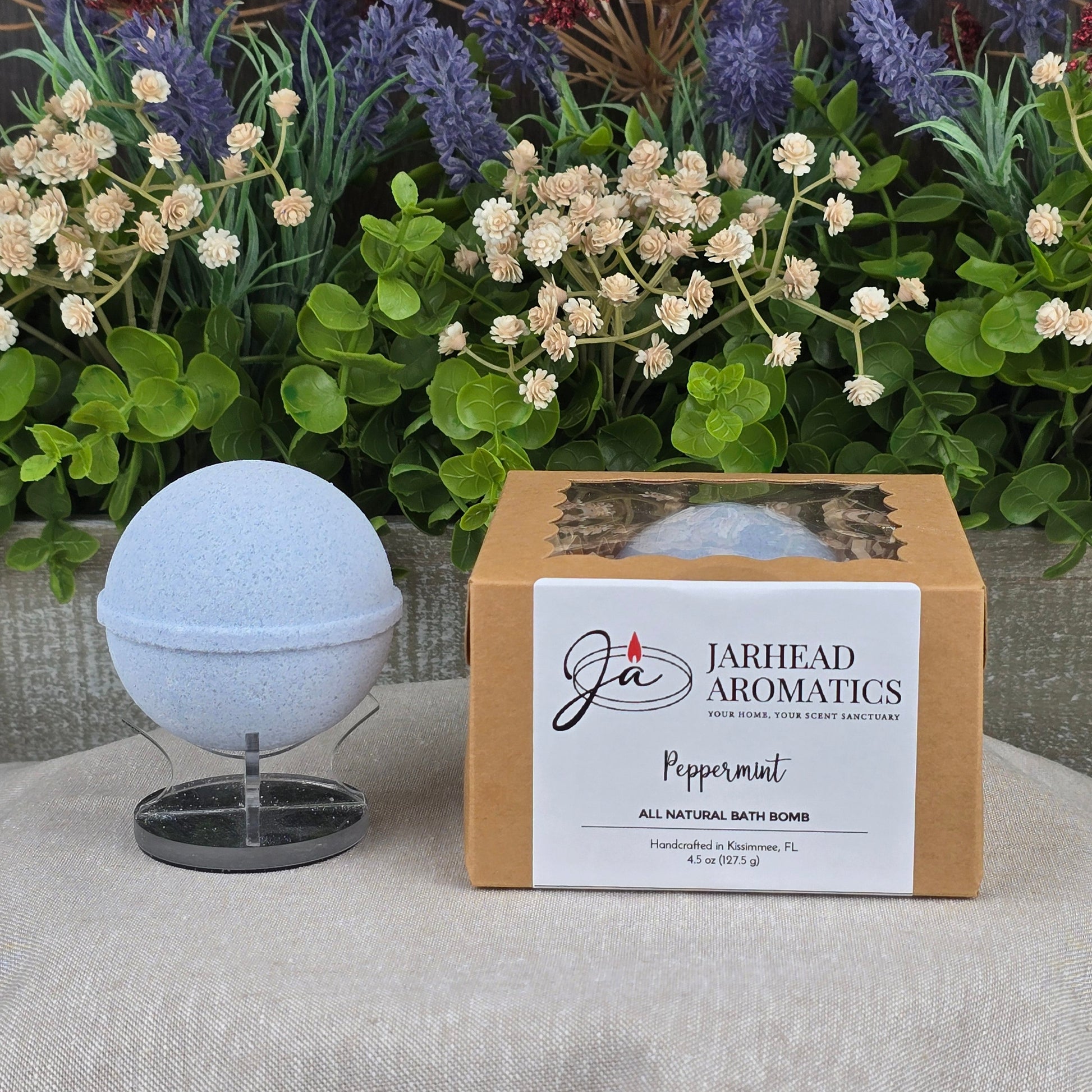 A light blue Jarhead Aromatics Peppermint Bath Bomb - All Natural sits on a stand beside its labeled box, displayed before vibrant artificial flowers.