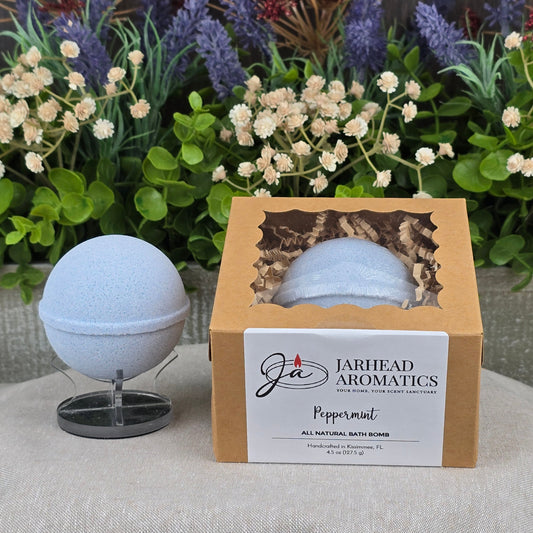 A light blue handmade bath bomb rests on a stand beside its box, labeled Jarhead Aromatics Peppermint Bath Bomb - All Natural. Colorful artificial flowers and greenery decorate the background.