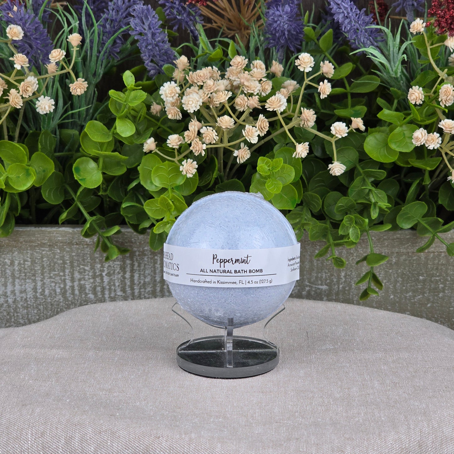 The Jarhead Aromatics Peppermint Bath Bomb - All Natural, a pale blue handmade bath bomb, sits on a small stand against lush green foliage and colorful artificial flowers atop a light gray textured surface.