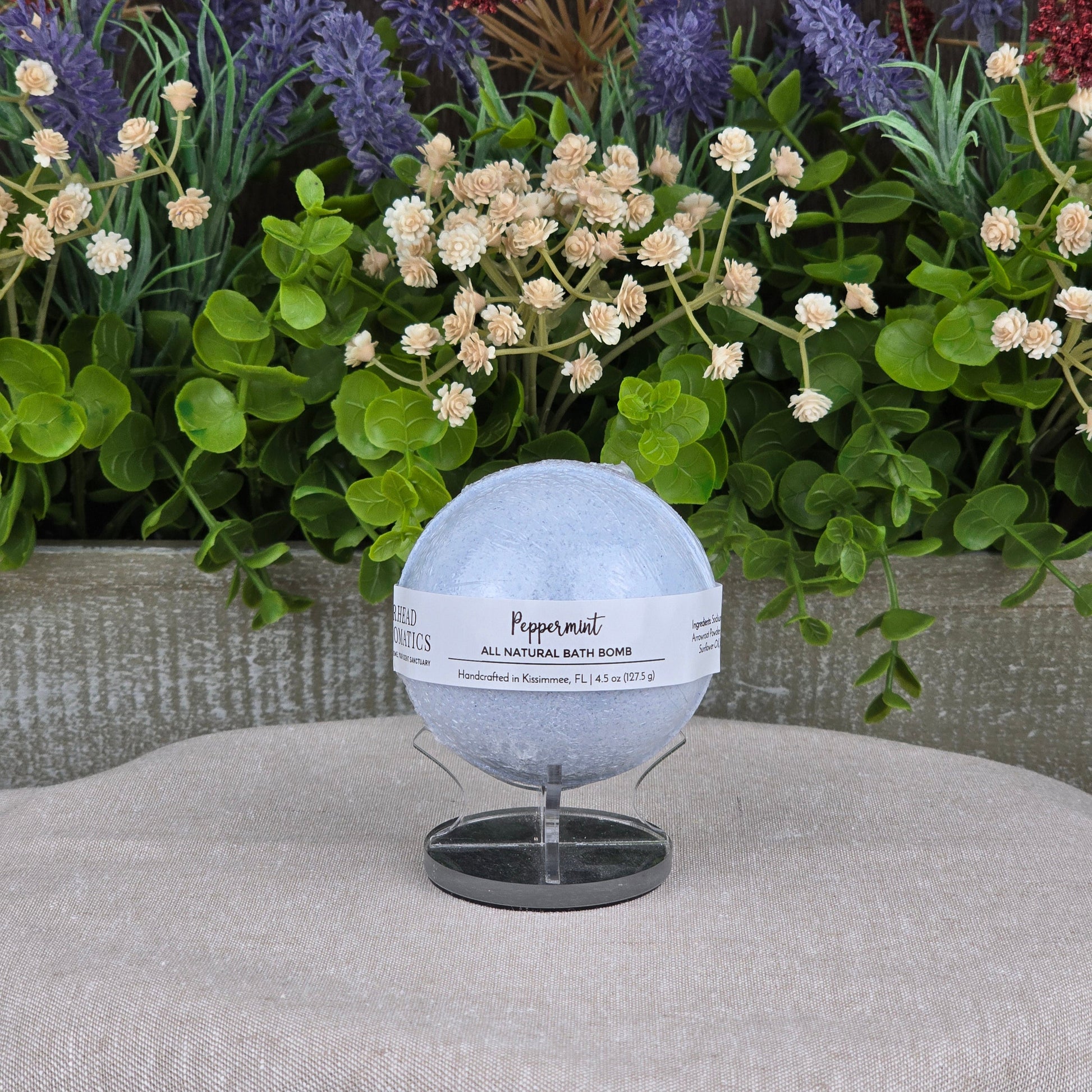The Jarhead Aromatics Peppermint Bath Bomb - All Natural, a pale blue handmade bath bomb, sits on a small stand against lush green foliage and colorful artificial flowers atop a light gray textured surface.