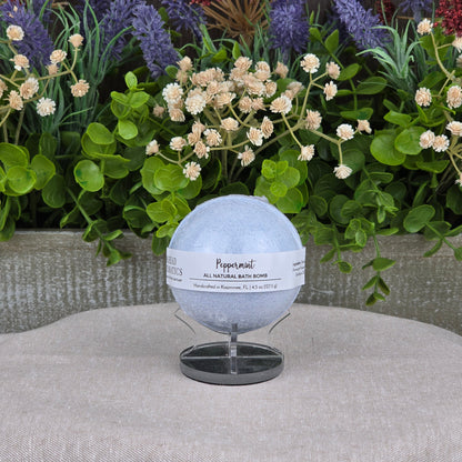 The Jarhead Aromatics Peppermint Bath Bomb - All Natural, a pale blue handmade bath bomb, sits on a small stand against lush green foliage and colorful artificial flowers atop a light gray textured surface.