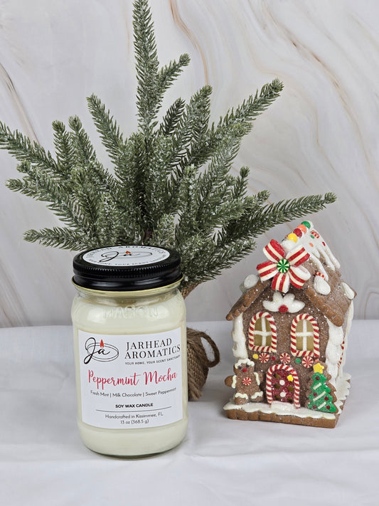 A Jarhead Aromatics Peppermint Mocha - Soy Wax Candle sits beside a candy-covered gingerbread house, complemented by dark chocolate details. A small pine branch arrangement completes the festive holiday backdrop.