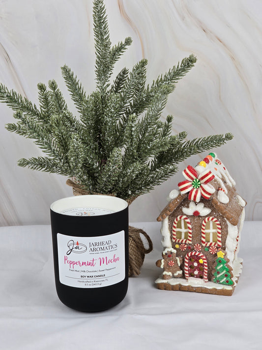 A black jar of Jarhead Aromatics Peppermint Mocha Soy Wax Candle sits near a small artificial pine tree and a gingerbread house with colorful candies, embodying holiday spirit. The background resembles marble, echoing rich dark chocolate swirls.