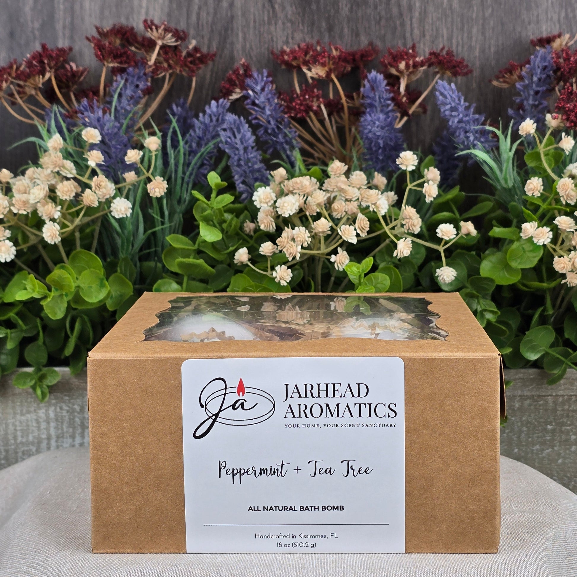 A Peppermint + Tea Tree Bath Bomb - All Natural by Jarhead Aromatics is nestled in a box, crafted with natural ingredients and showcased against lush greenery and purple flowers.