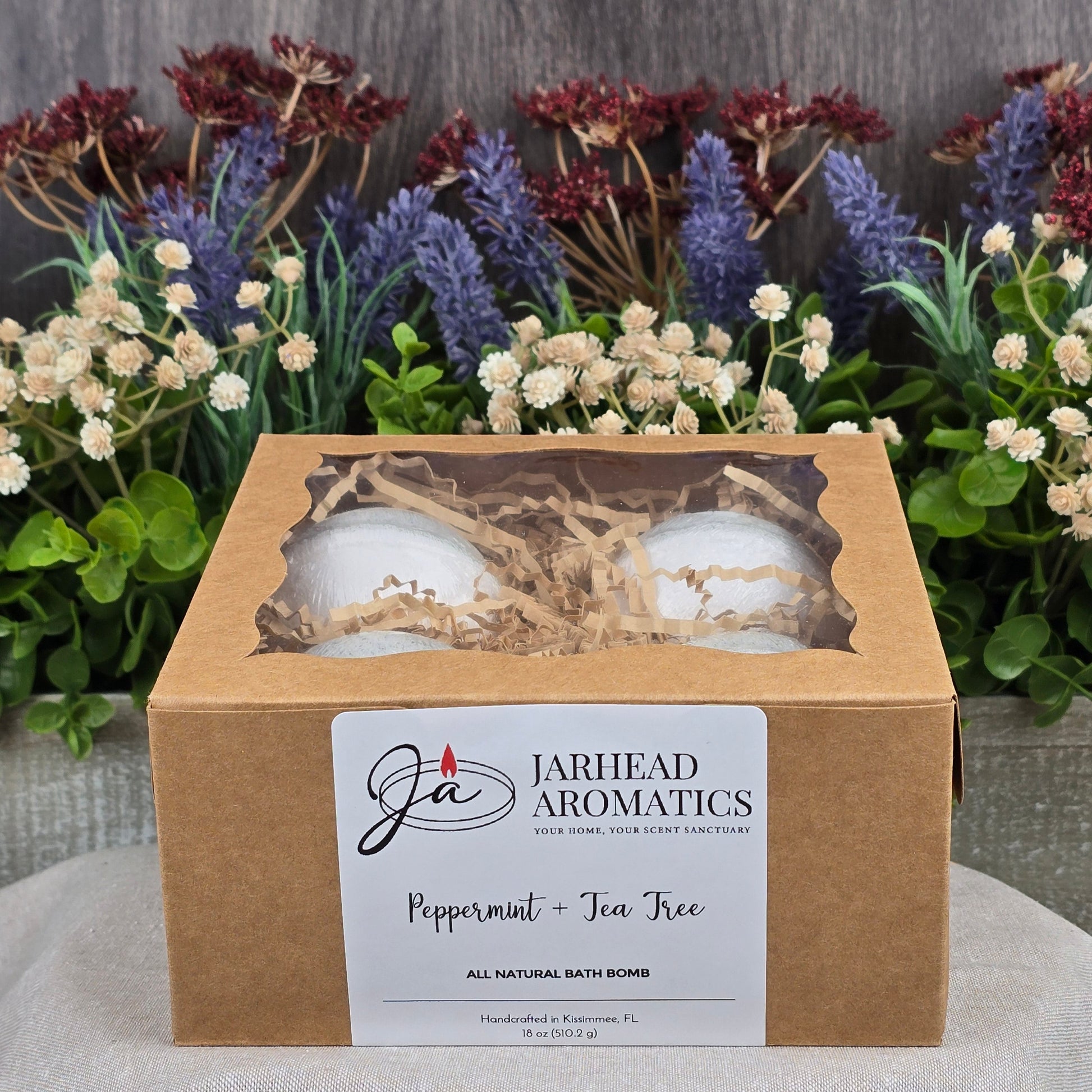 A brown cardboard box from Jarhead Aromatics contains four Peppermint + Tea Tree Bath Bombs - All Natural, nestled in shredded paper, against vibrant flowers. These handmade bath bombs promise a refreshing escape.