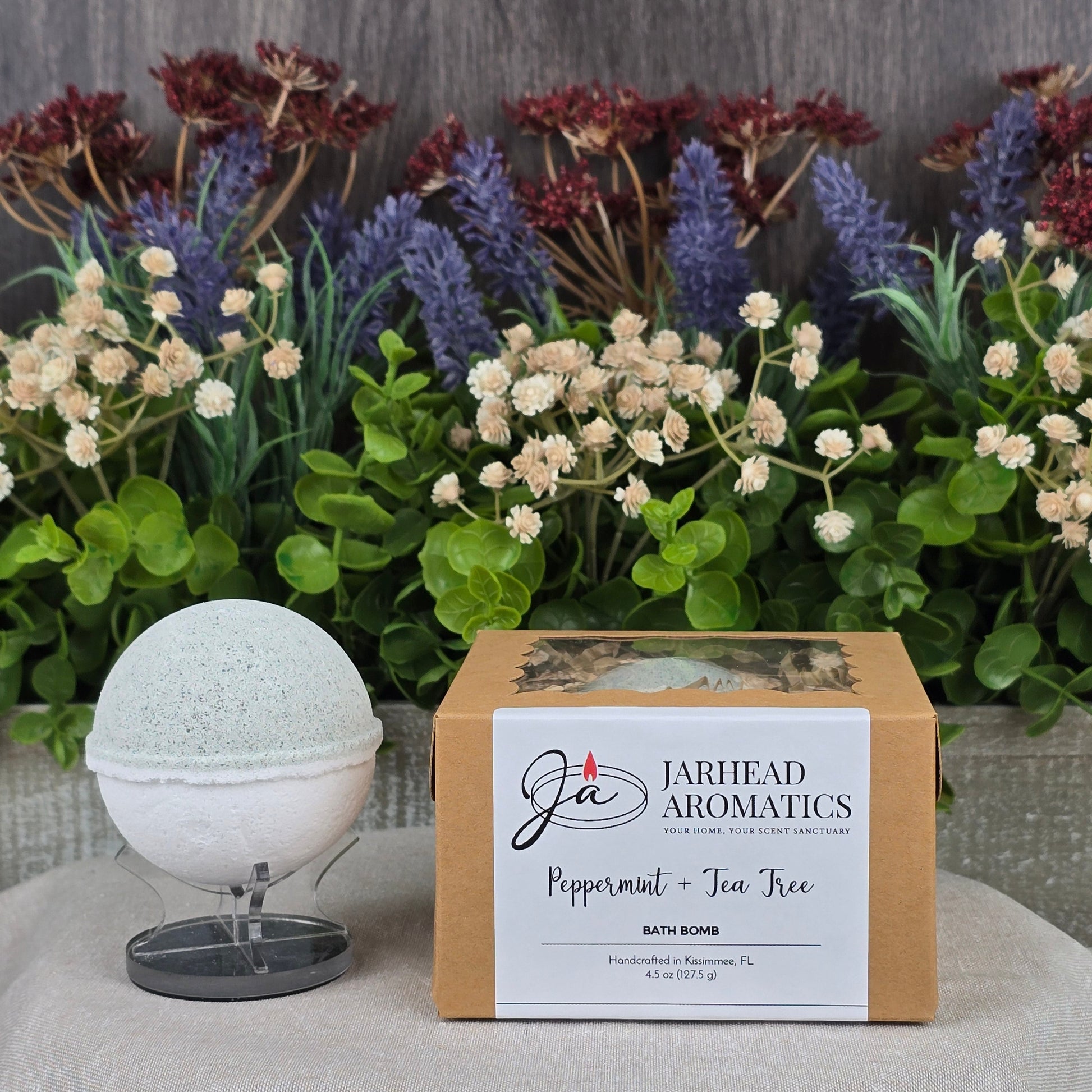 The Peppermint + Tea Tree Bath Bomb from Jarhead Aromatics, boasting all-natural ingredients, is showcased with its box against a textured gray background. A lush arrangement of purple, white, and brown flowers accents the invigorating handmade bath bomb.