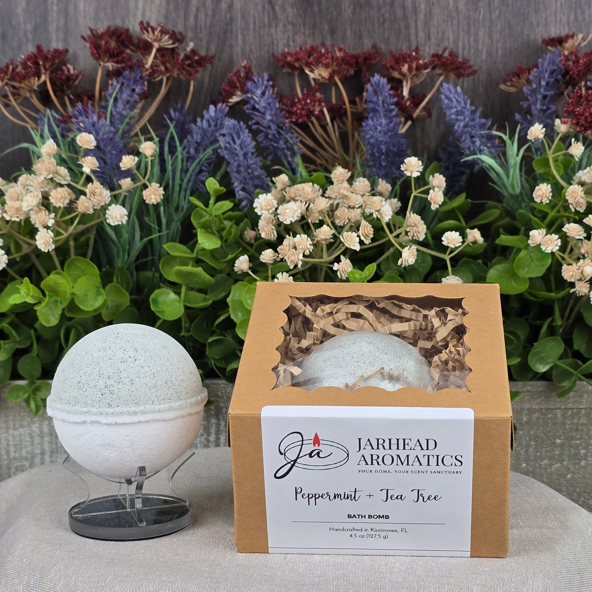 The Peppermint + Tea Tree Bath Bomb by Jarhead Aromatics, handmade and all-natural, sits elegantly in its box against vibrant greenery and colorful flowers.
