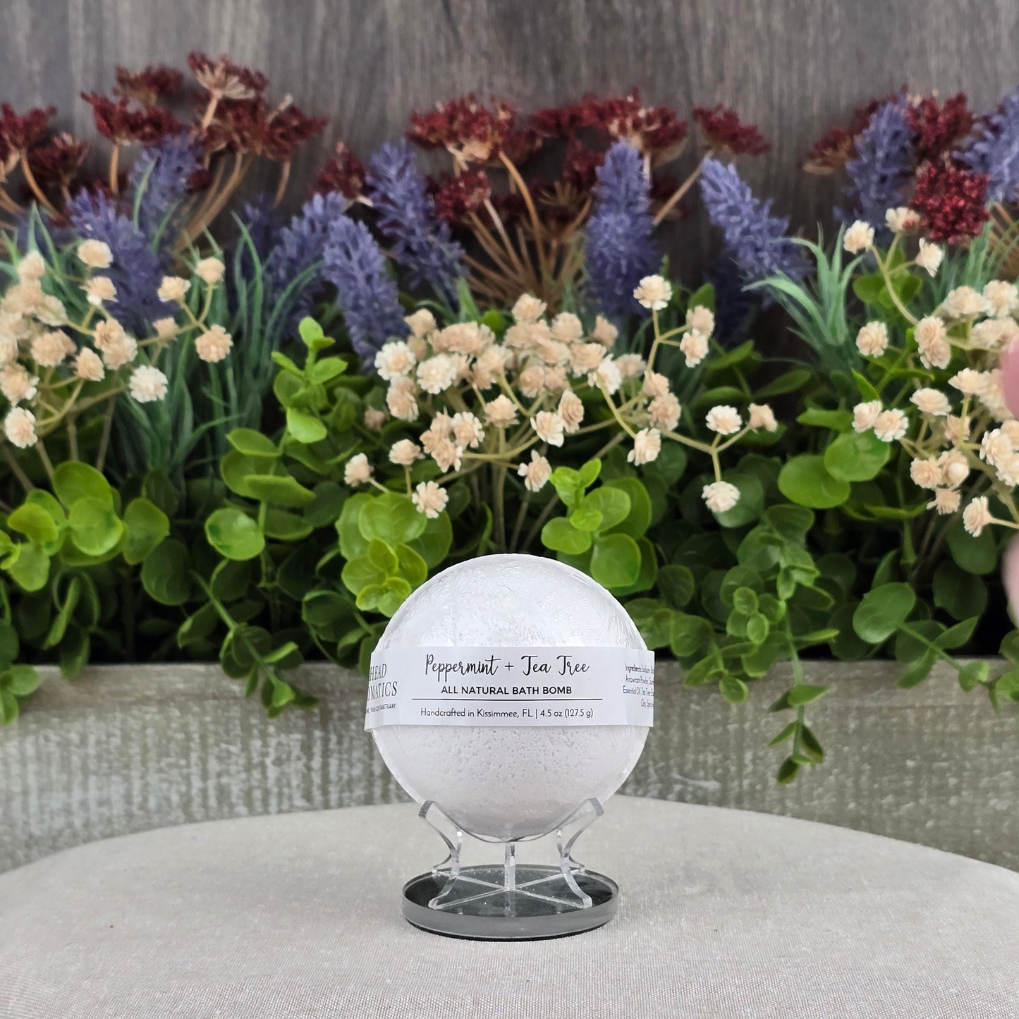 The Jarhead Aromatics Peppermint + Tea Tree Bath Bomb – All Natural is displayed on a stand over a beige cloth, accented by faux greenery and vibrant flowers to emphasize its natural ingredients.