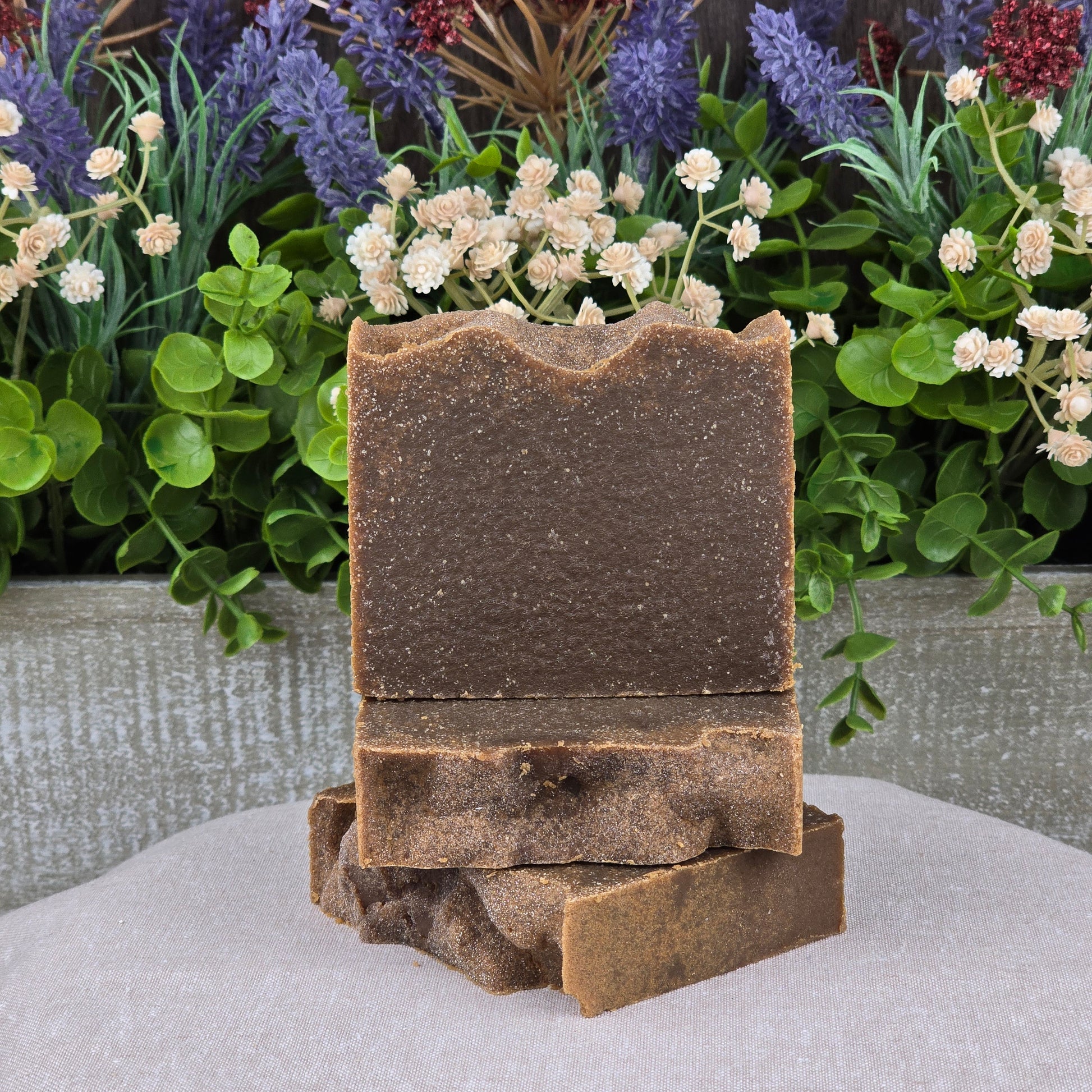 Two brown Pine Tar Handmade Natural Artisan Soap bars by Jarhead Aromatics are stacked on a beige cloth, accented by green leaves and purple and white flowers. Their rustic texture highlights their woodsy, artisan quality with cocoa butter & essential oils.