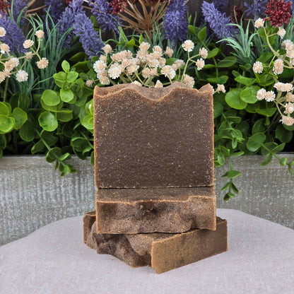 Two brown Pine Tar Handmade Natural Artisan Soap bars by Jarhead Aromatics are stacked on a beige cloth, accented by green leaves and purple and white flowers. Their rustic texture highlights their woodsy, artisan quality with cocoa butter & essential oils.
