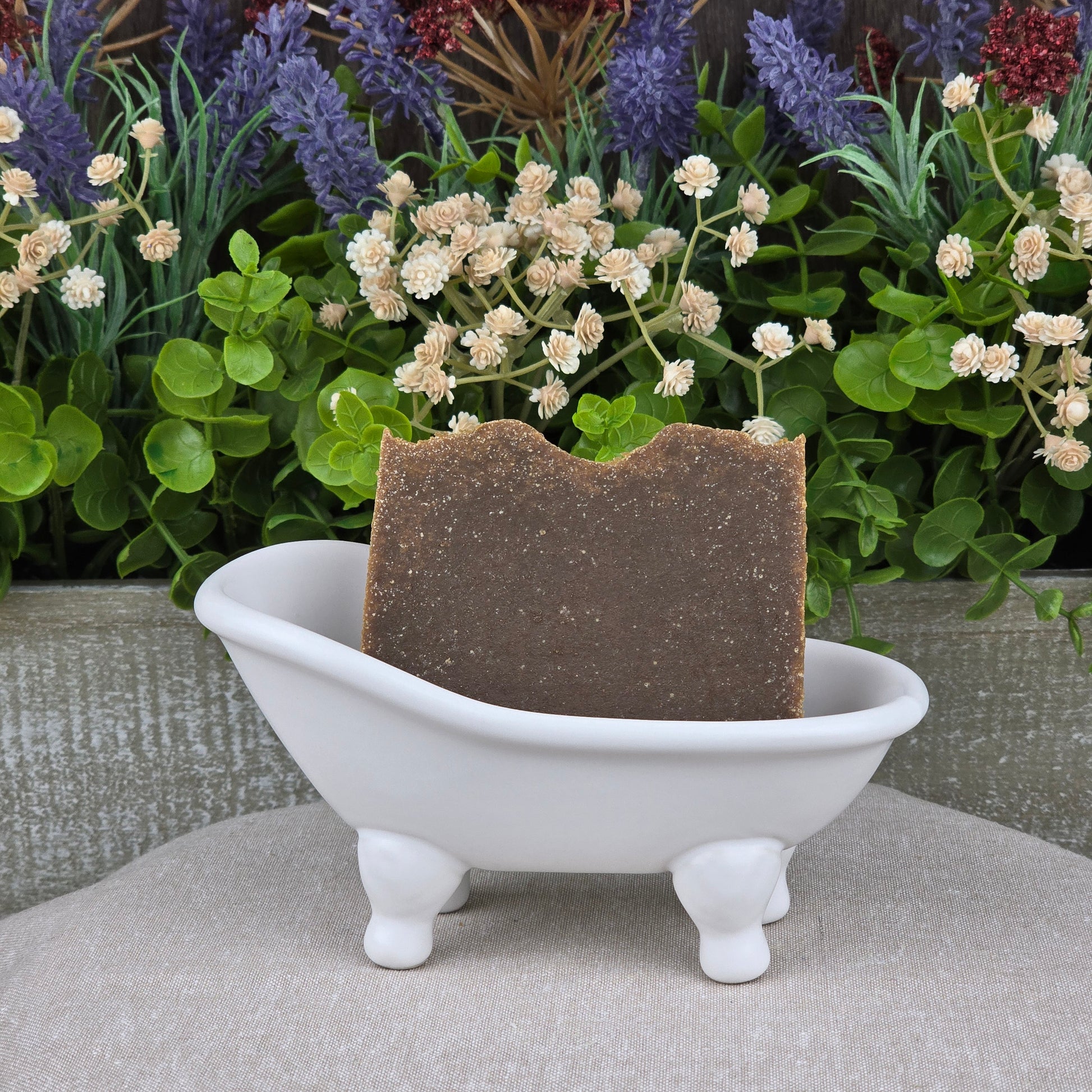 A Pine Tar Handmade Natural Artisan Soap by Jarhead Aromatics rests in a white clawfoot bathtub soap dish, with green and purple artificial flowers in the background.