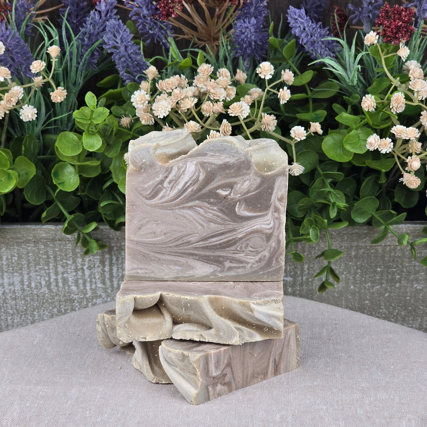 Two Porter + Coffee Handmade Craft Beer Artisan Soaps by Jarhead Aromatics, featuring a swirled, natural pattern, are stacked on a beige surface with artificial leaves and colorful flowers in the background.
