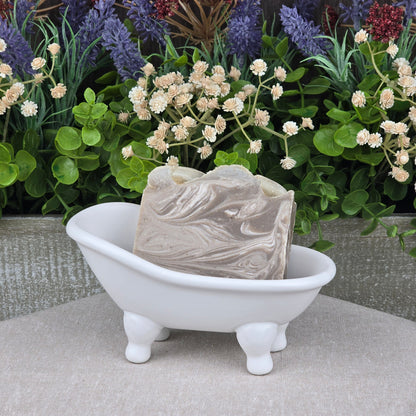 The Jarhead Aromatics Porter + Coffee Handmade Craft Beer Artisan Soap with coffee exfoliant and porter beer sits in a white bathtub-shaped dish, with artificial green leaves and colorful flowers on a wooden background.